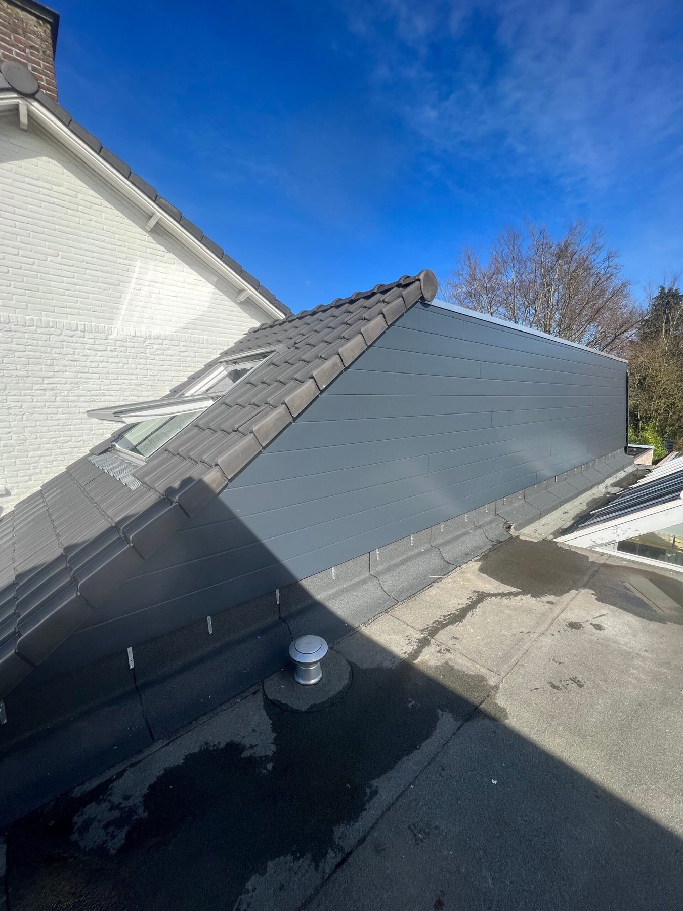 Zijkant van een nieuwe dakopbouw met grijze gevelbekleding, antracieten dakpannen en een dakraam, onder een blauwe lucht